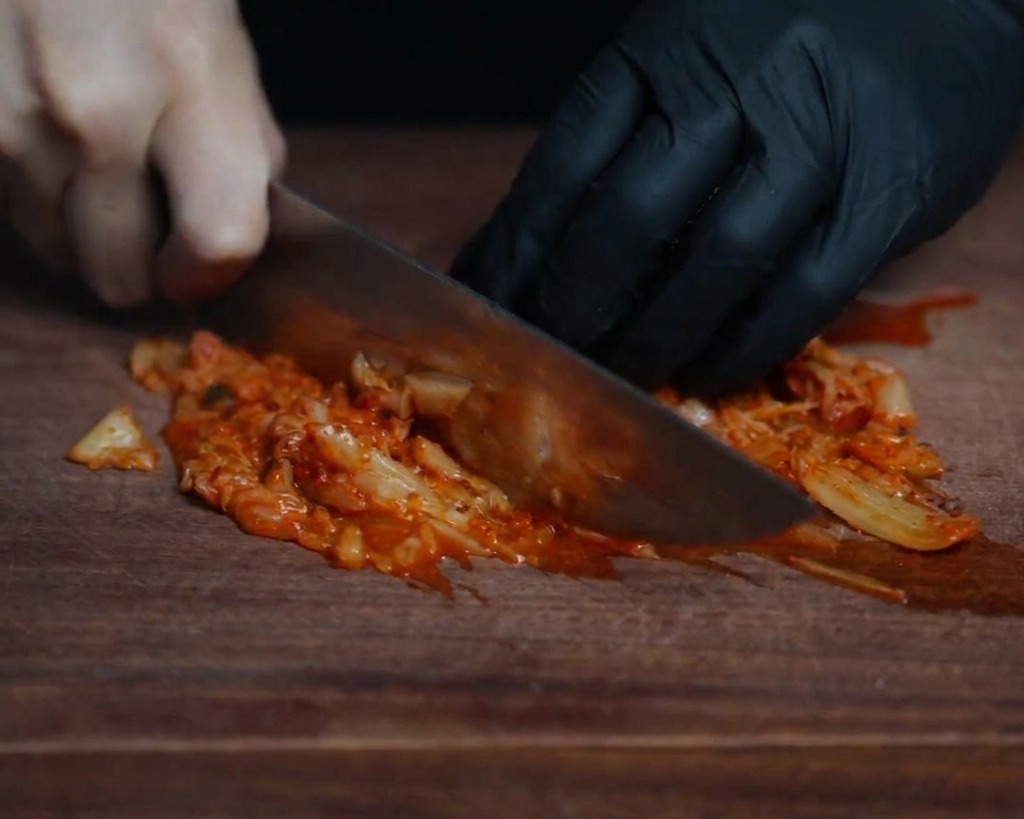 Primer plano de un chef picando finamente kimchi rojo brillante sobre una tabla de madera oscura con un cuchillo afilado.