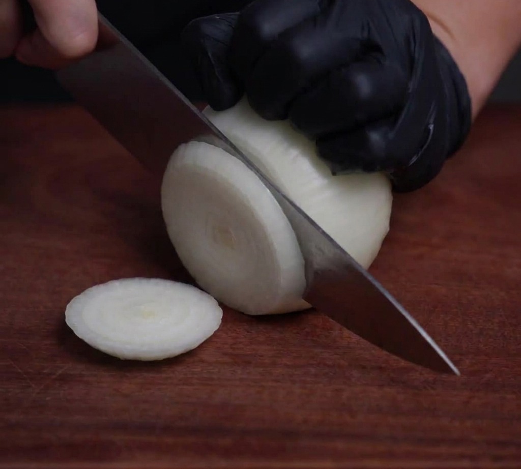 Una cebolla blanca cortada en aros finos y translúcidos sobre una superficie de madera usando un cuchillo de chef profesional.
