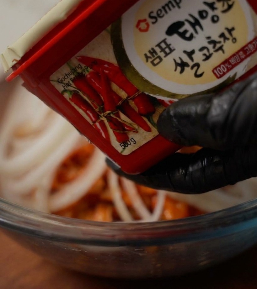 Una mano con guante negro sujetando un envase de pasta de chile rojo sobre un bol para añadir una cucharada de pasta.
