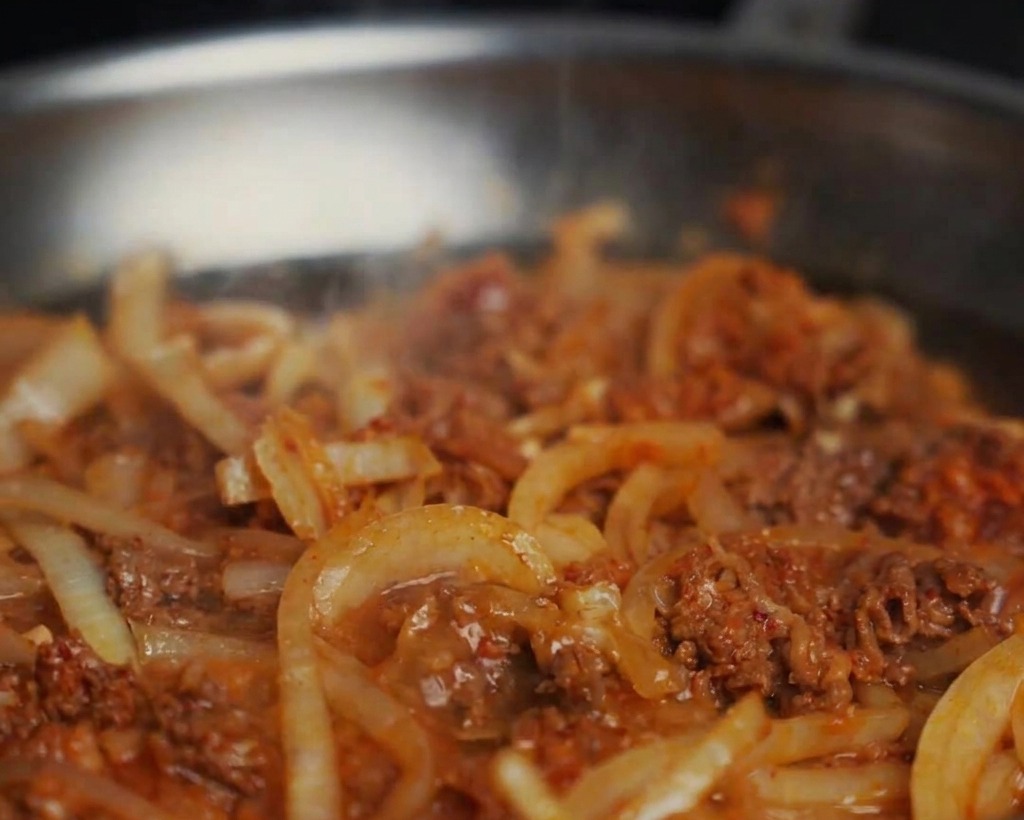 Agua añadiéndose a una sartén con ternera y cebollas salteadas, creando una salsa burbujeante.