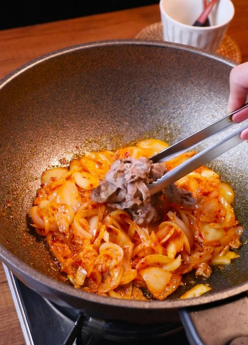 Pinzas devolviendo las láminas de ternera a la sartén con el kimchi y la cebolla.