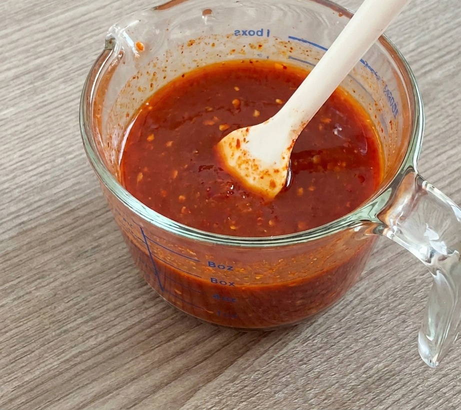 Una espátula de silicona blanca removiendo una salsa espesa y roja en una jarra de cristal.