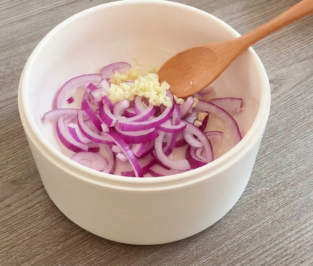 Aros de cebolla roja y ajo picado siendo salteados con una cuchara de madera en una sartén blanca.