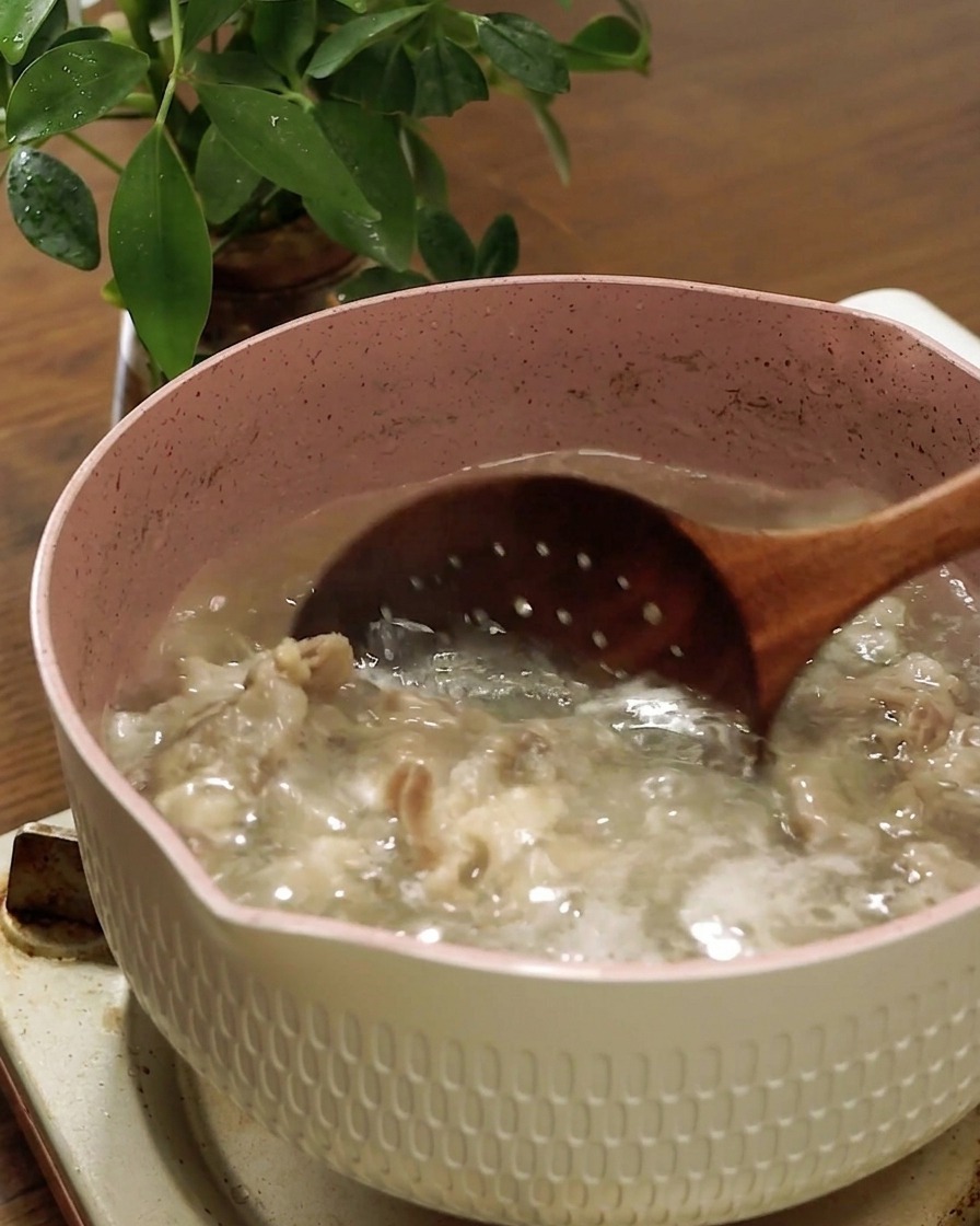 Una espumadera de madera retirando la ternera cocida de una olla con agua hirviendo.