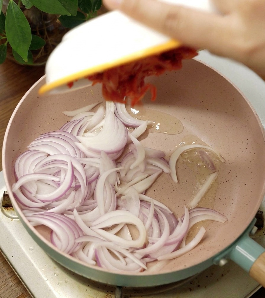 Sartén cerámica rosa con cebolla morada en juliana y una porción de kimchi picante que se añade desde un bol blanco.
