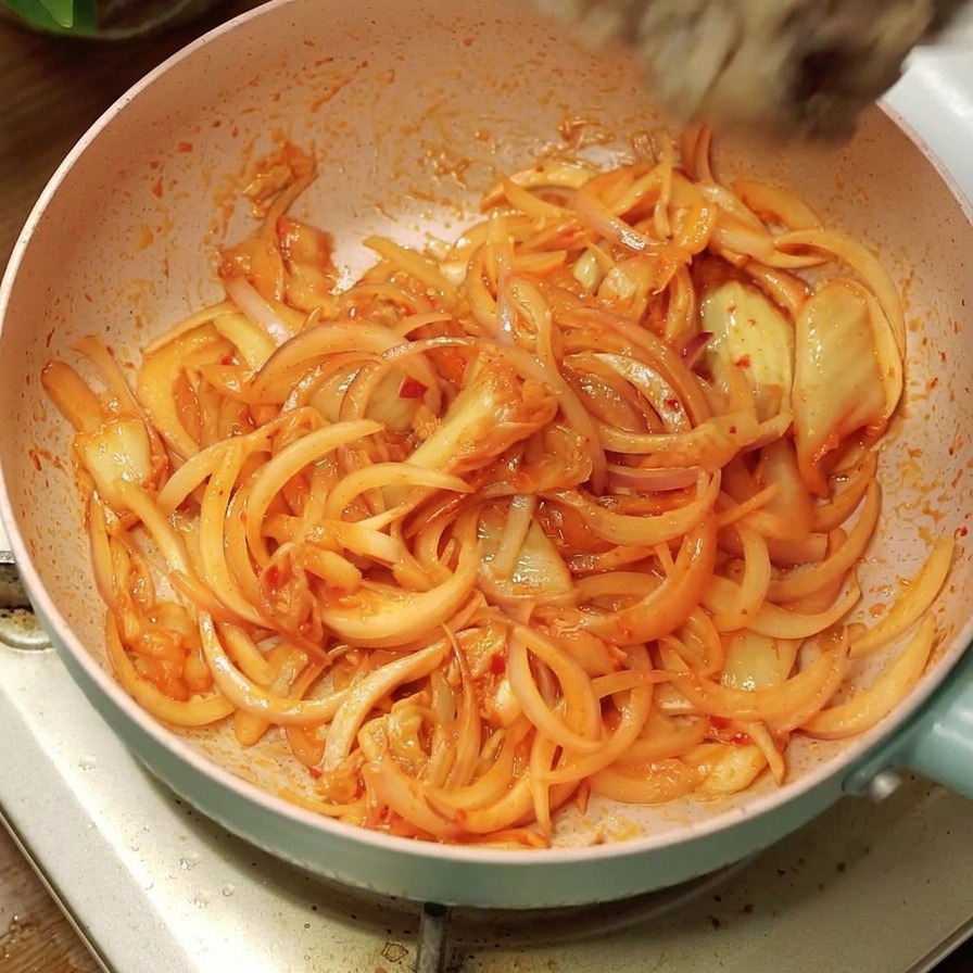 Láminas de carne cocida vertiéndose desde un bol blanco a una sartén rosa con cebollas y kimchi salteados.