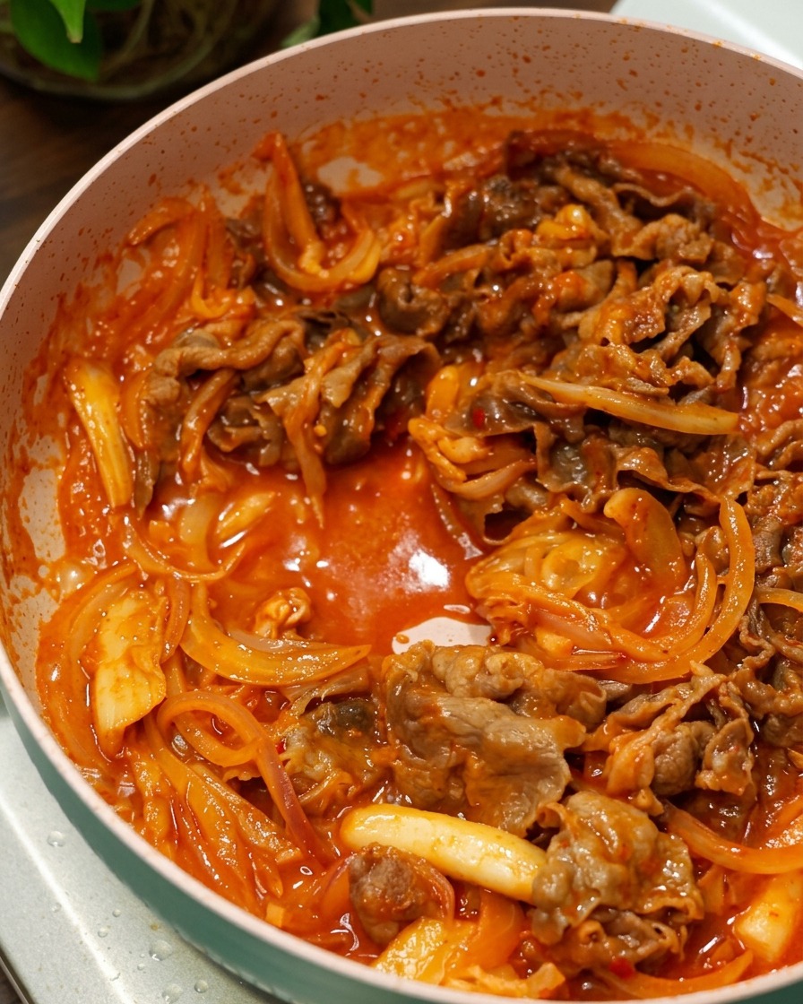 Ternera y cebollas salteándose en una salsa de chile coreana roja y burbujeante dentro de una sartén rosa.