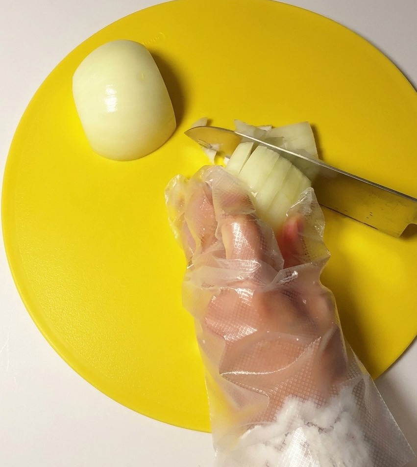 Mano con guante de plástico picando una cebolla blanca con un cuchillo de acero sobre una tabla de cortar amarilla.
