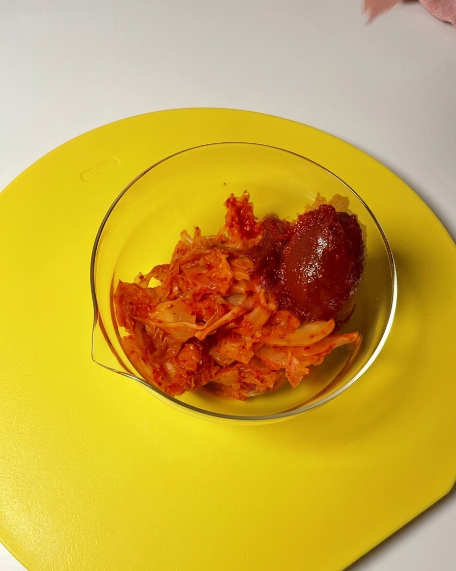 Añadiendo una cucharada de pasta gochujang y chile en polvo a un bol de cristal con kimchi troceado.