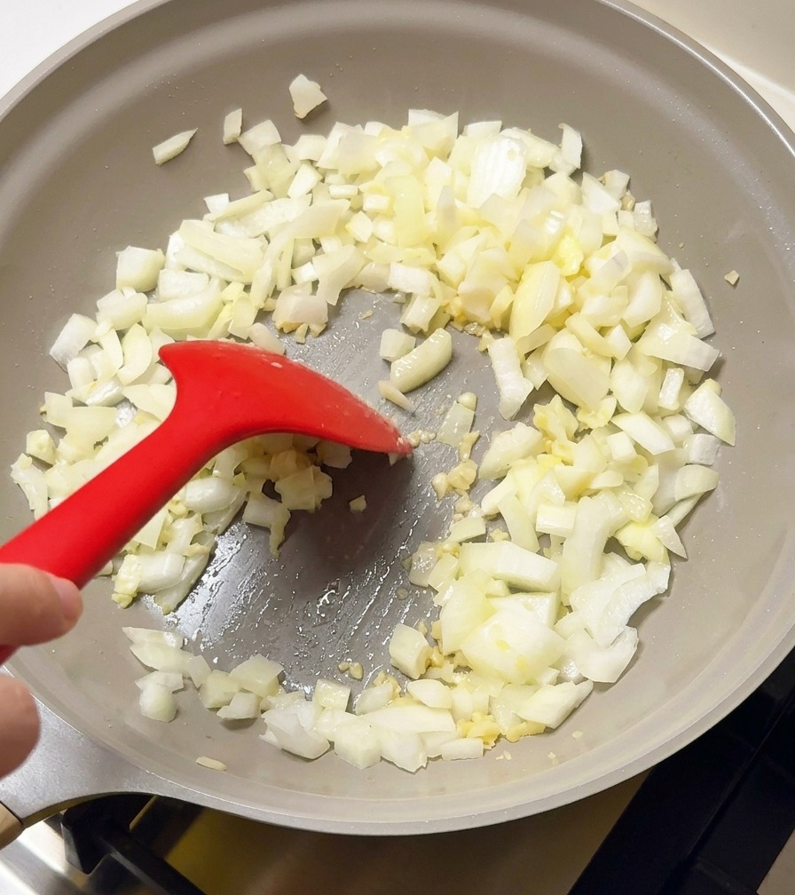 Cebolla blanca y ajo picados sofriéndose en una sartén gris con una espátula de silicona roja.