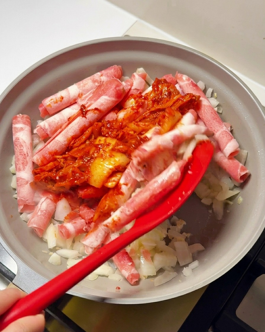 Espátula roja añadiendo la salsa de kimchi y gochujang sobre la ternera y la cebolla en la sartén.