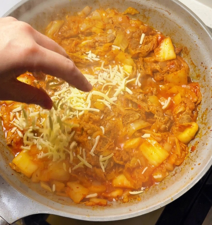Mano espolvoreando una generosa cantidad de mozzarella rallada sobre el salteado picante de kimchi y ternera.