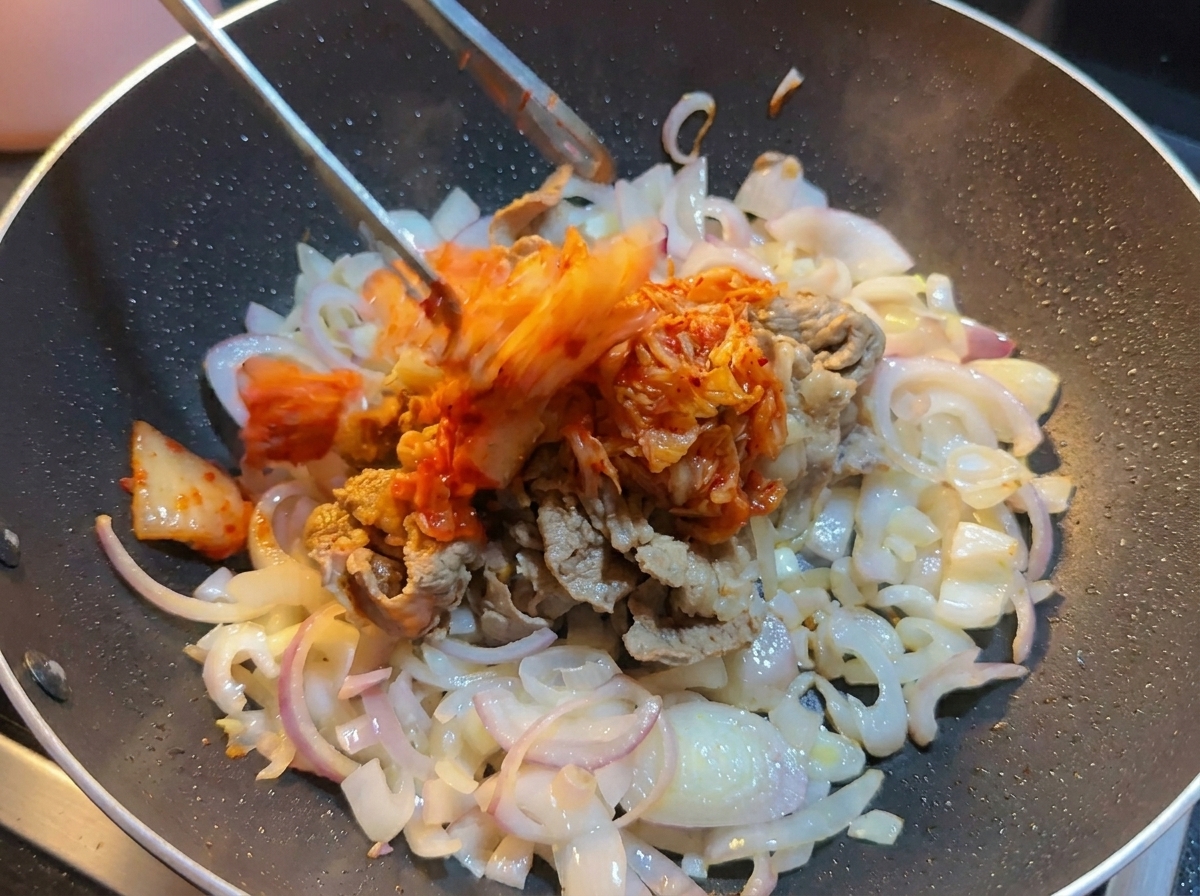 Kimchi de col picado siendo añadido con pinzas a un wok que contiene ternera y cebollas.