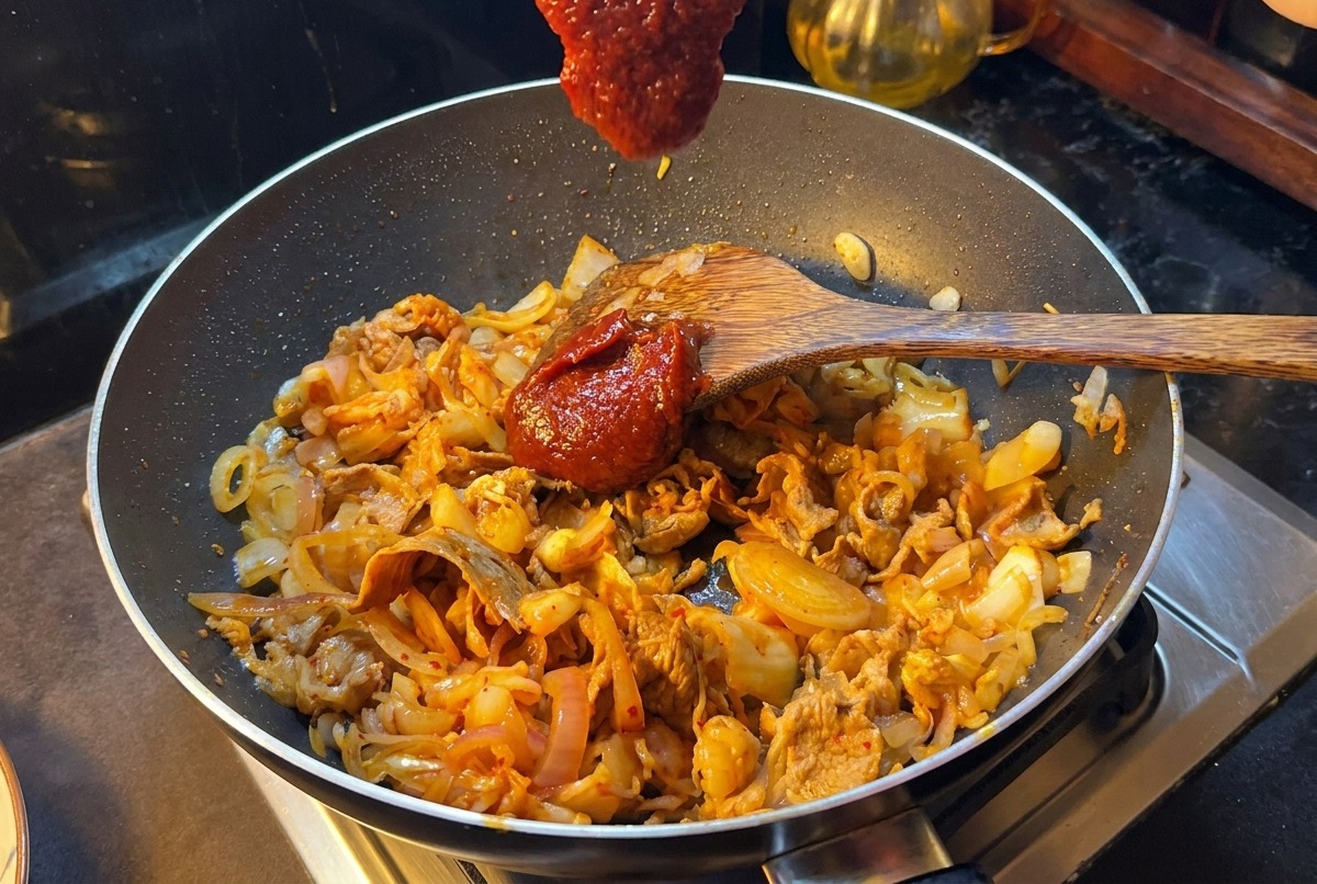 Una espátula de madera con una porción de pasta de chile coreana roja sobre la sartén con ternera y kimchi.