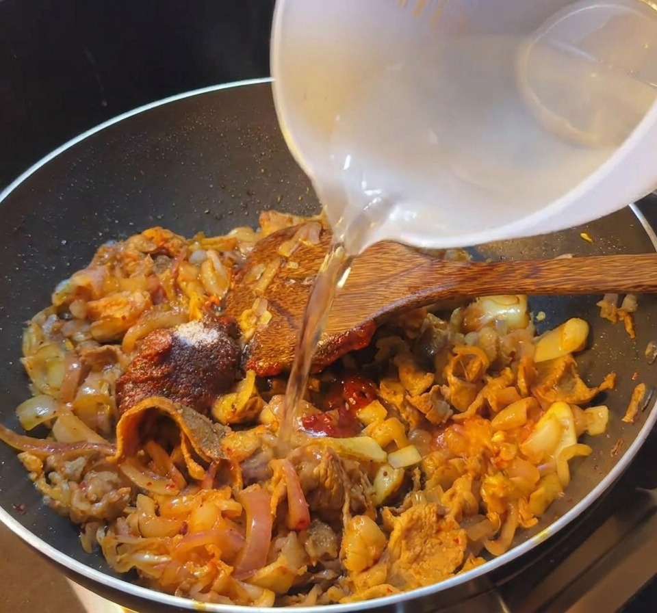 Persona vertiendo agua desde un bol blanco en una sartén con ternera y salsa de kimchi picante.