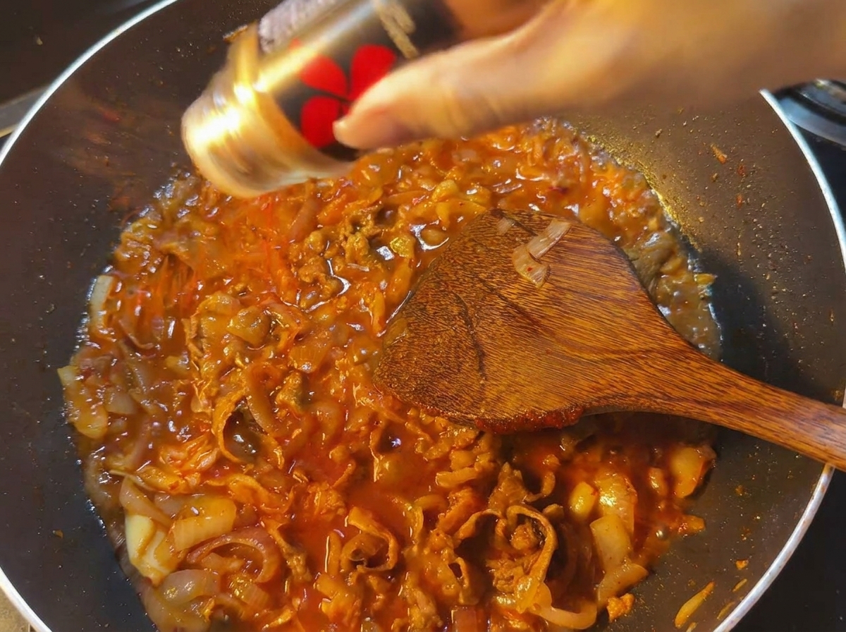 Polvo de chile rojo cayendo sobre una sartén con salsa burbujeante, ternera y cebollas.