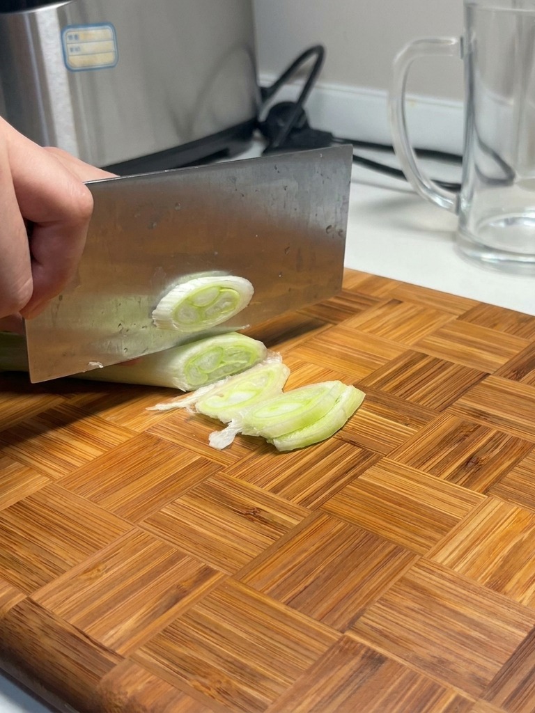 Un cuchillo de cocina cortando un puerro grueso en rodajas blancas y verdes sobre una tabla de madera.