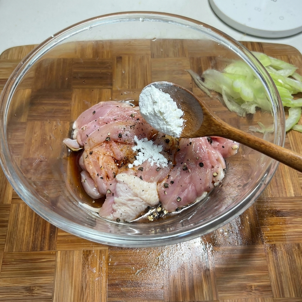 Una cuchara de madera añadiendo maicena blanca a un bol de cristal con trozos de pollo crudo sazonados.