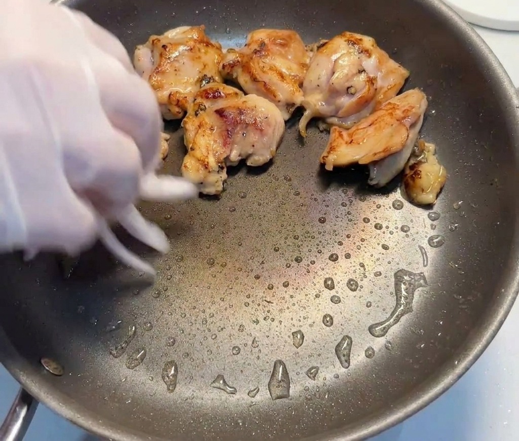 Rodajas de cebolla roja siendo añadidas por una mano a una sartén que contiene trozos de contramuslo de pollo sellados.