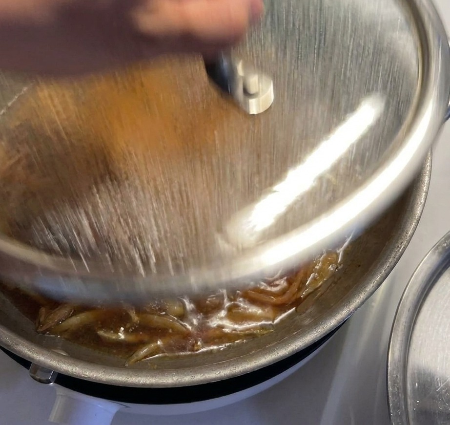 Una mano colocando una tapa de cristal sobre una sartén donde el pollo y las cebollas hierven en una salsa oscura.