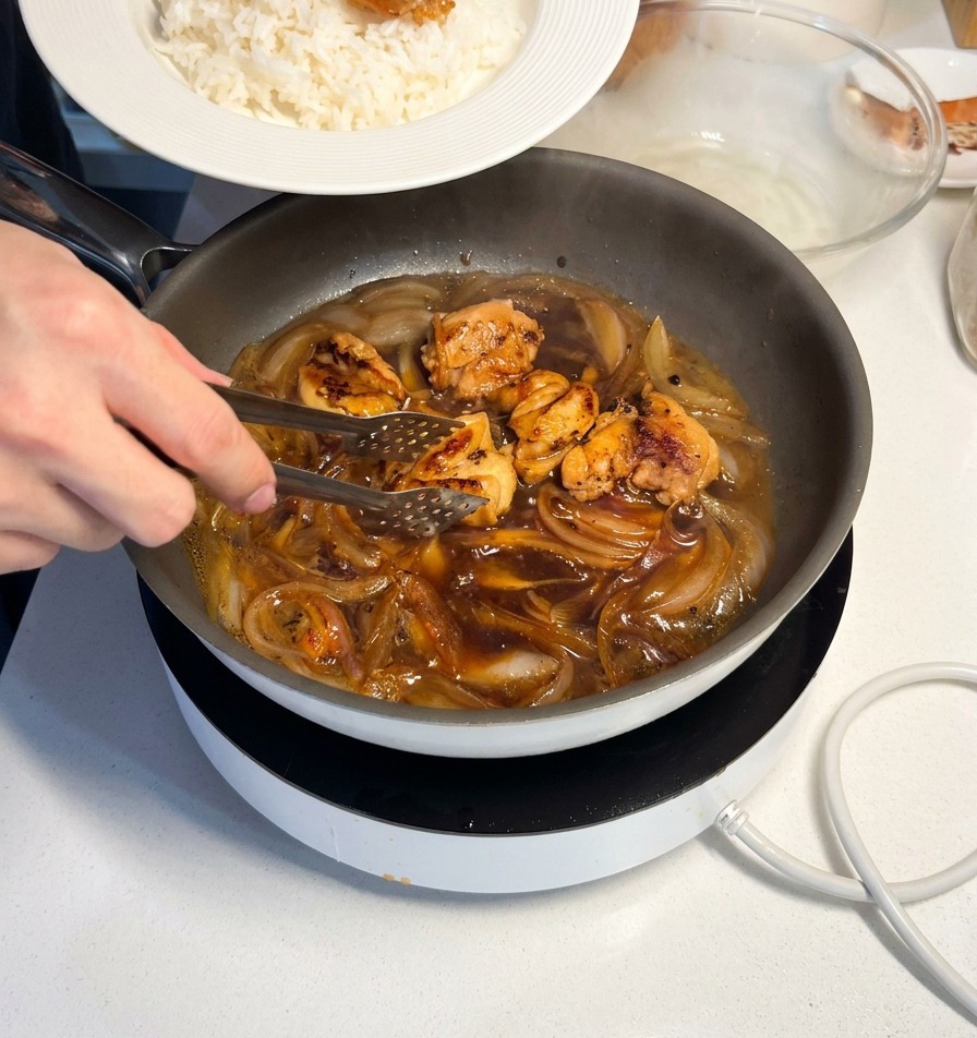 Pinzas de metal colocando un trozo de pollo glaseado sobre una montaña de arroz blanco en un bol de cerámica.