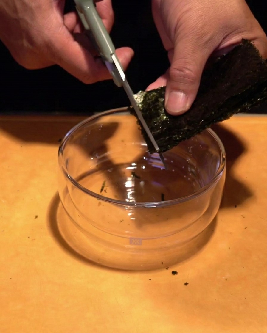 Manos usando tijeras de cocina para cortar una hoja de alga nori en tiras finas sobre un cuenco de cristal.