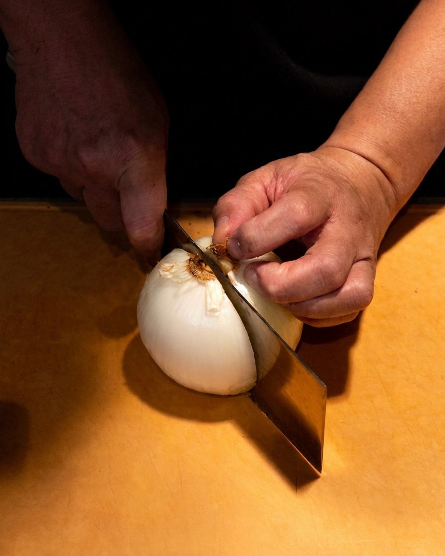 Primer plano de unas manos cortando con cuidado una cebolla blanca en gajos finos sobre una tabla de madera.
