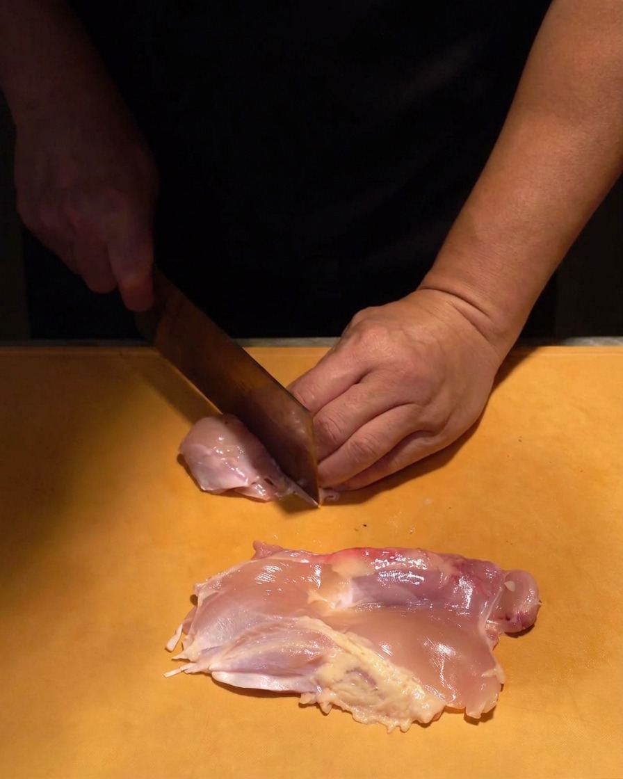 Carne de contramuslo de pollo cruda siendo cortada en trozos uniformes con un cuchillo afilado sobre una tabla naranja.