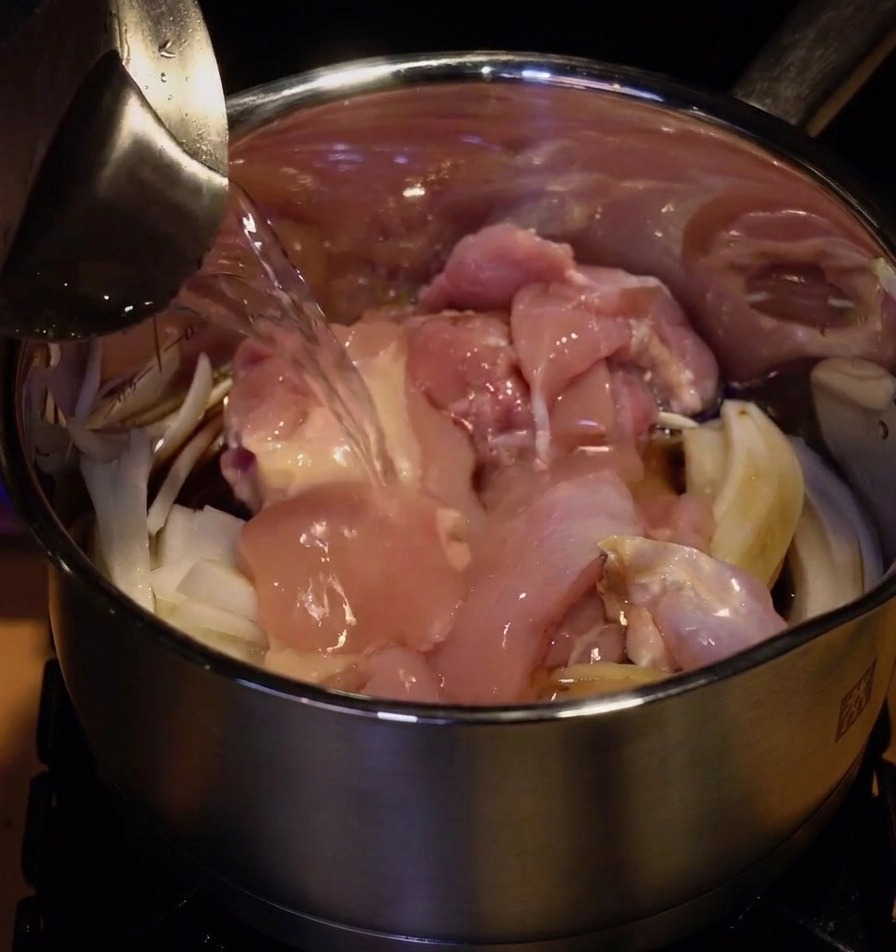 Agua siendo vertida desde una jarra metálica en la olla con el pollo crudo y la cebolla troceada.