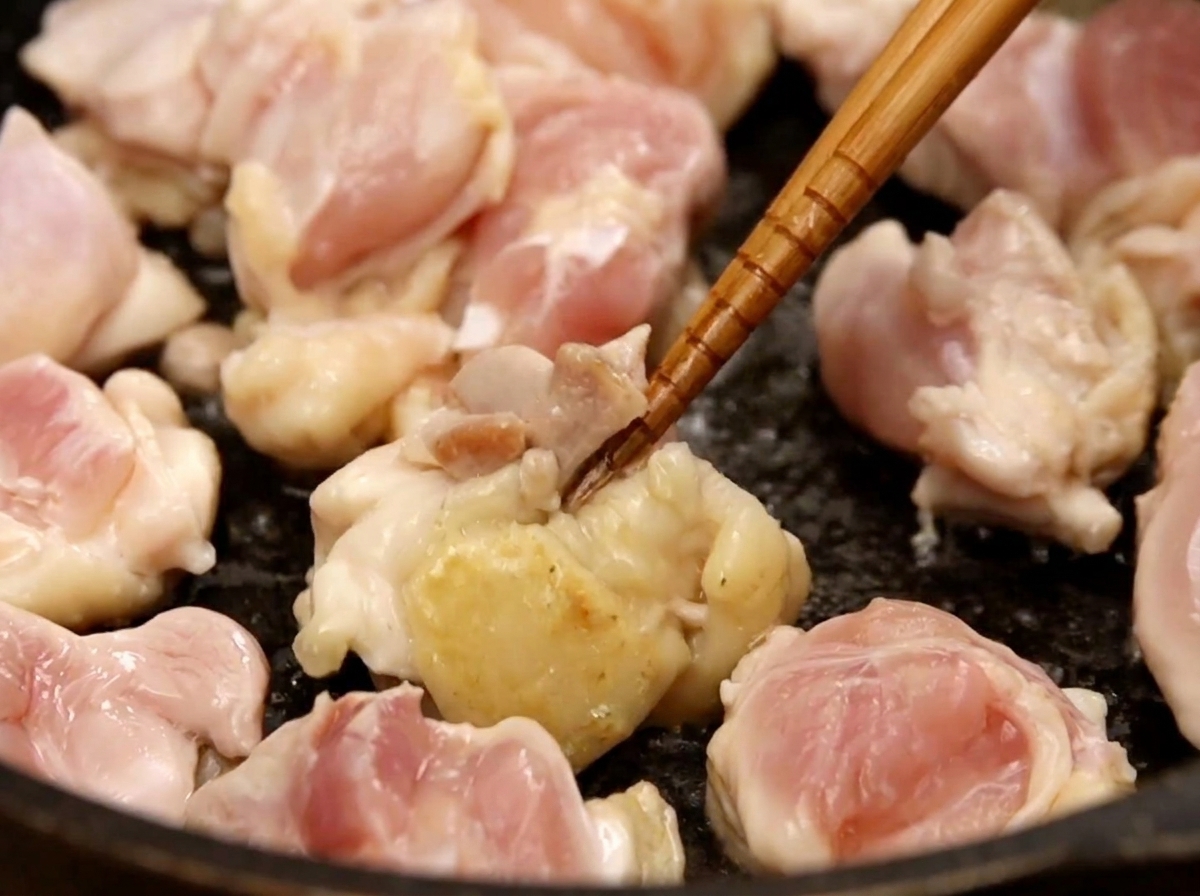 Dados de pollo salteándose en una sartén oscura con palillos, mostrando algunas piezas empezando a dorarse.