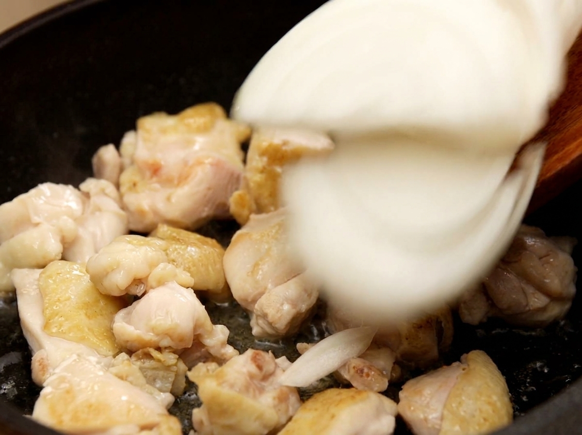 Añadiendo cebolla blanca en tiras a la sartén con el pollo salteado y mezclándolo todo.