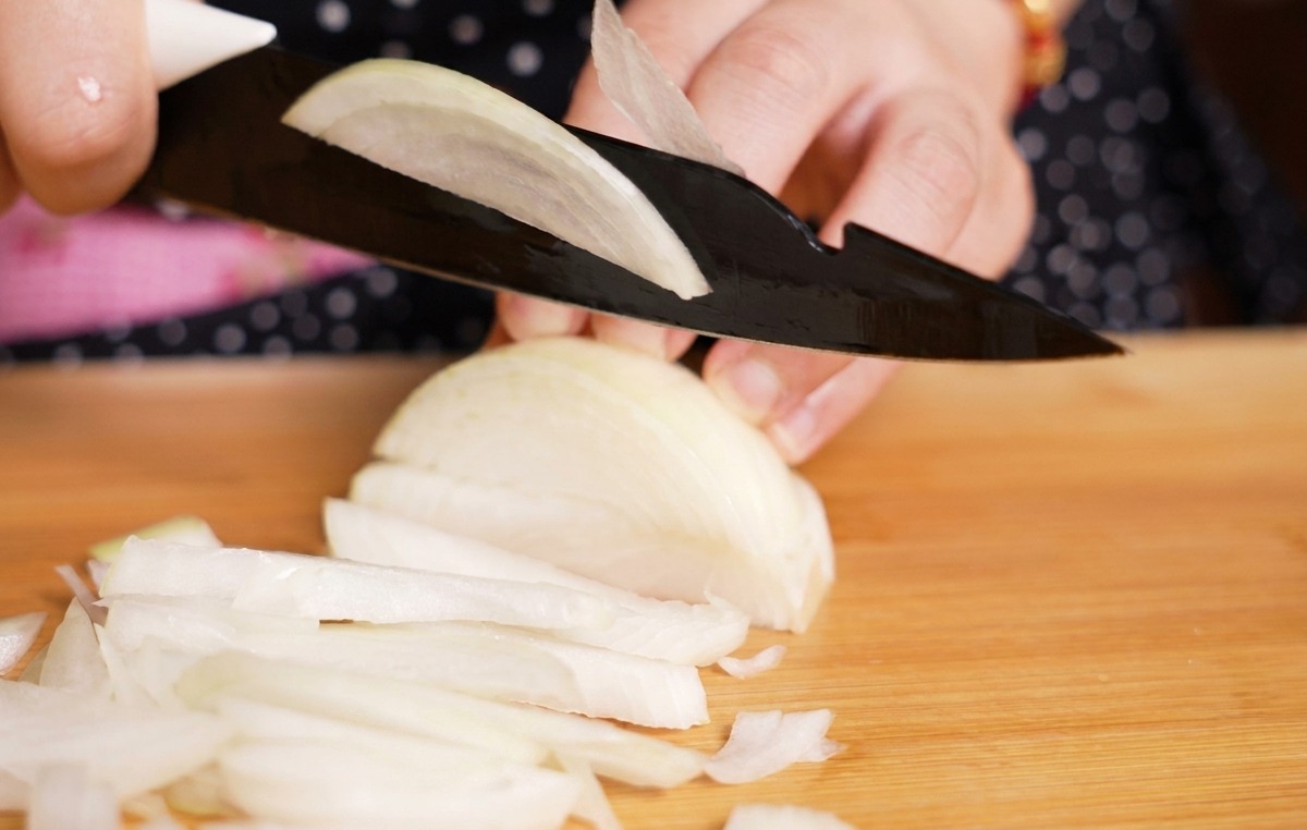 Una persona usando un cuchillo negro para cortar una cebolla blanca en tiras finas sobre una tabla de madera clara.