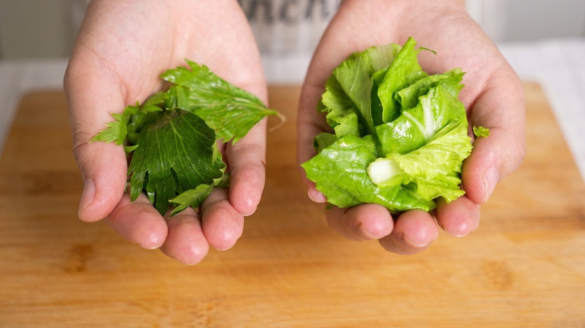 Dos manos sosteniendo puñados de hojas de apio picadas y bok choy pequeño sobre una tabla de bambú.