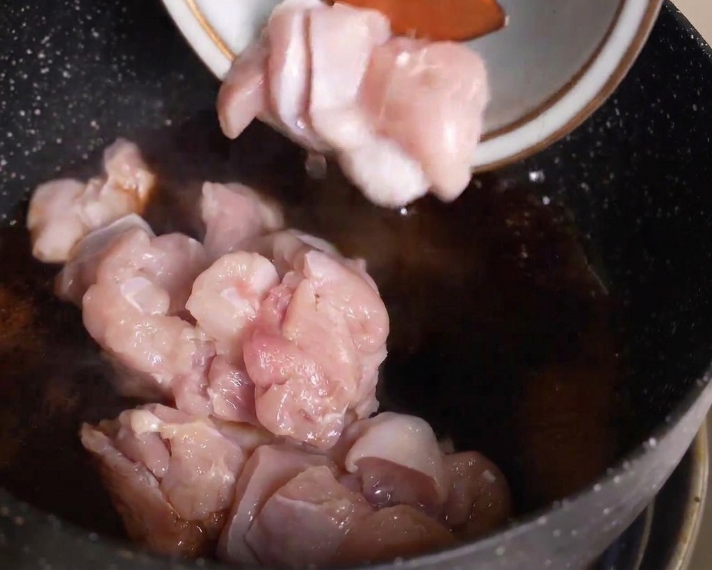 Trozos de pollo crudo siendo añadidos desde un bol a una sartén con salsa oscura hirviendo.