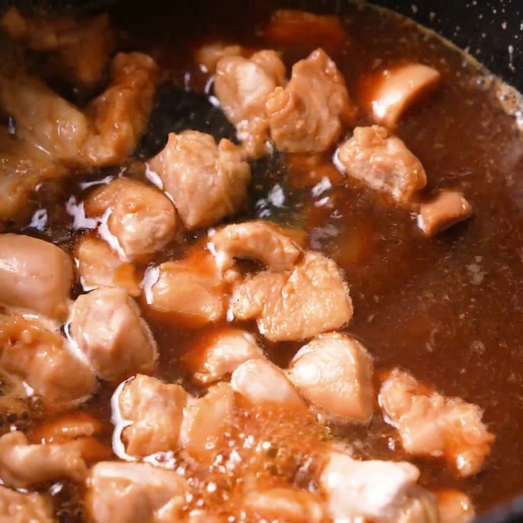 Trozos de pollo cocinándose en un caldo dashi de color marrón burbujeante dentro de una sartén oscura.