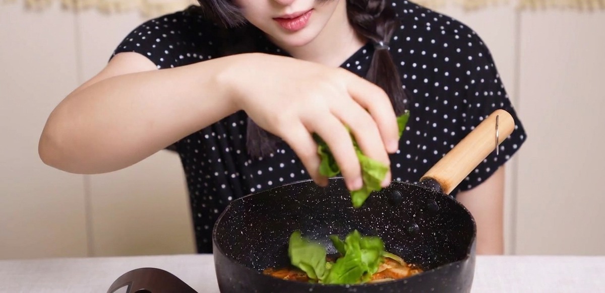 Verduras frescas siendo esparcidas a mano en una sartén con pollo, cebollas y huevos en salsa dashi.