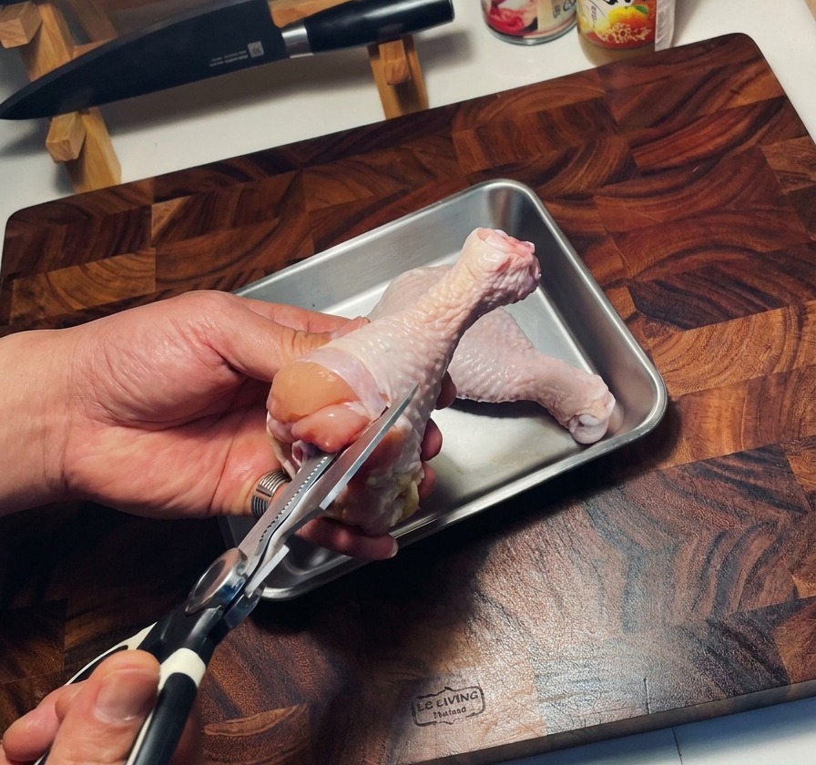 Unas tijeras de cocina quitando el hueso de un muslo de pollo crudo sobre una bandeja de metal.