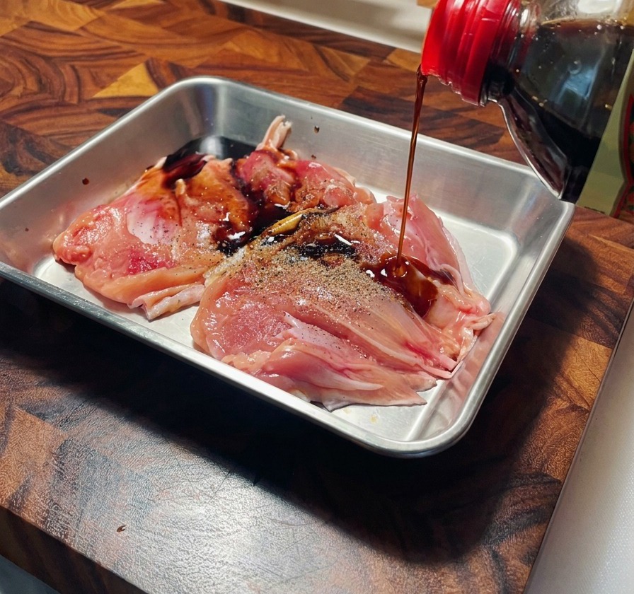 Salsa de soja vertiéndose desde una botella sobre muslos de pollo crudos con pimienta en una bandeja de acero inoxidable.