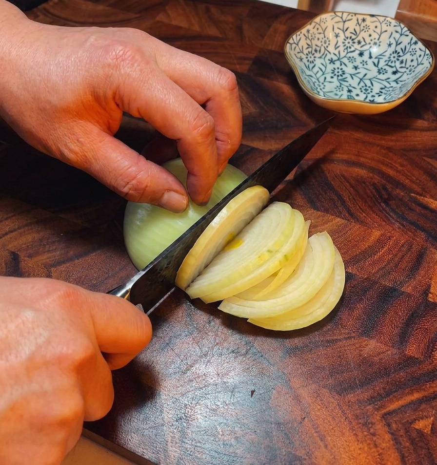 Las manos de un chef usando un cuchillo para cortar una cebolla amarilla en tiras finas sobre una tabla de cortar de madera oscura.