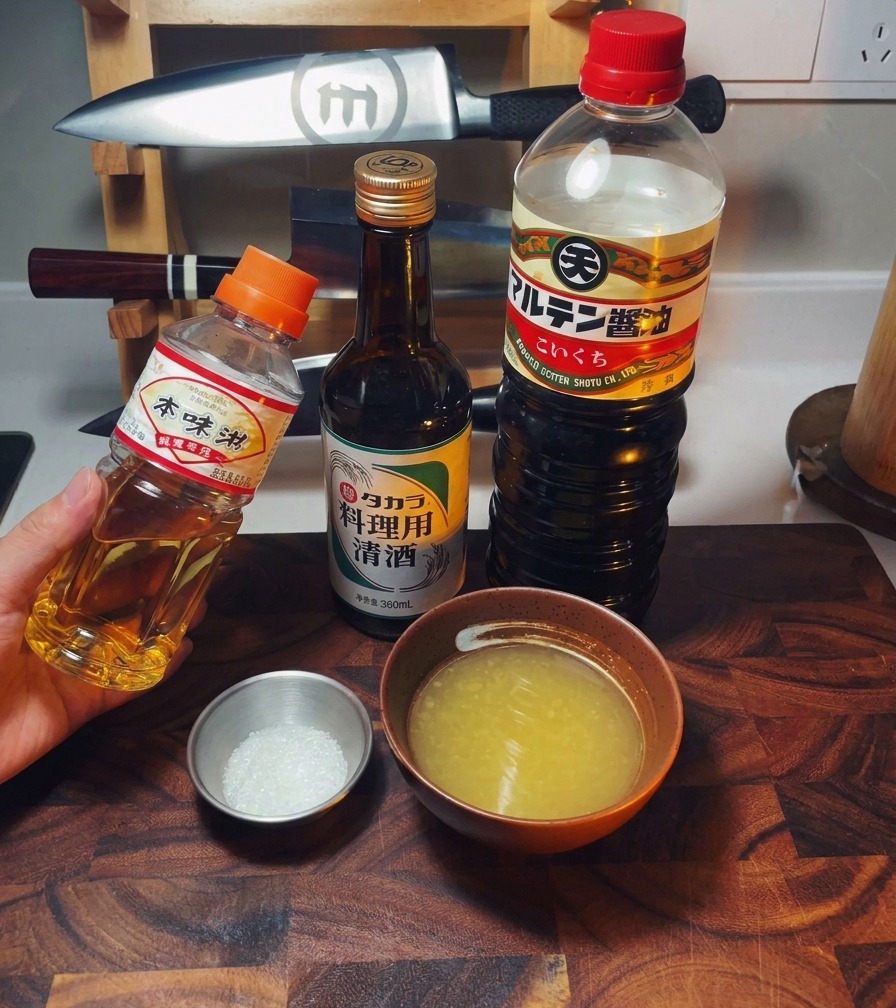Un arreglo de botellas de mirin, sake y salsa de soja junto a un tazón de caldo de pollo y azúcar.