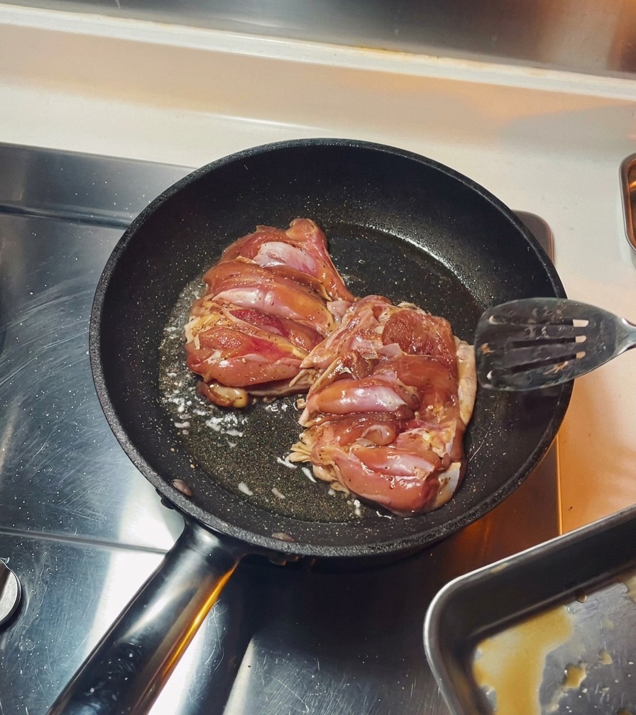Dos muslos de pollo sazonados friéndose con la piel hacia abajo en una sartén negra hasta dorarse.