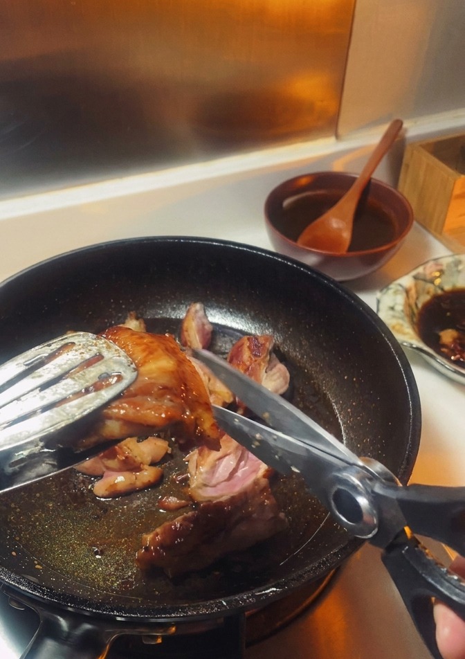 Unas tijeras de cocina cortando un muslo de pollo dorado sostenido por pinzas de metal dentro de una sartén.