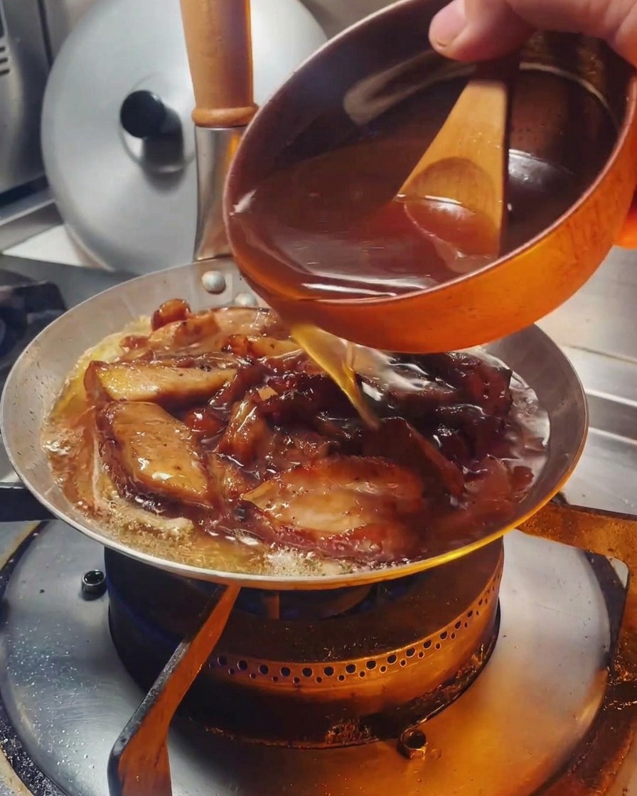 Una salsa marrón sabrosa vertiéndose desde un pequeño cuenco con mango de madera en una sartén que contiene pollo y cebollas.