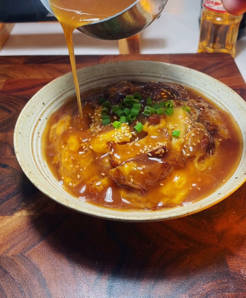 Una vista cercana de una mano usando un pequeño cucharón de metal para verter una salsa sabrosa y dorada sobre un cuenco de pollo y huevo adornado con cebolletas verdes.
