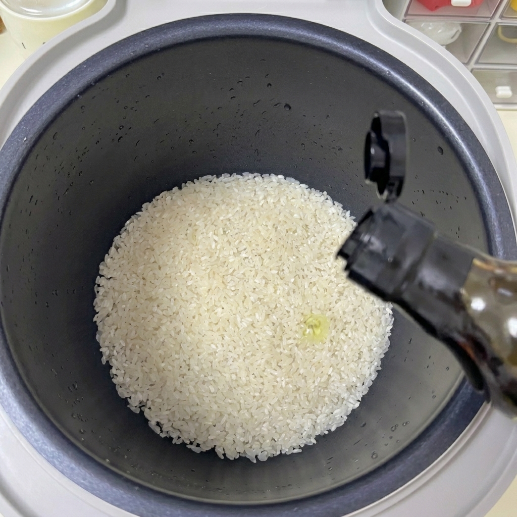 Una mano vertiendo un chorro de aceite transparente desde una botella oscura en una olla arrocera llena de arroz blanco.