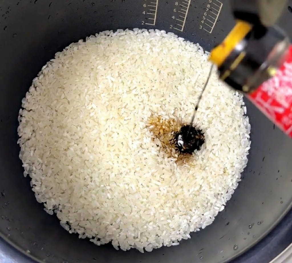 Un chorro de salsa de ostras oscura y espesa vertiéndose en una olla arrocera con arroz blanco lavado.