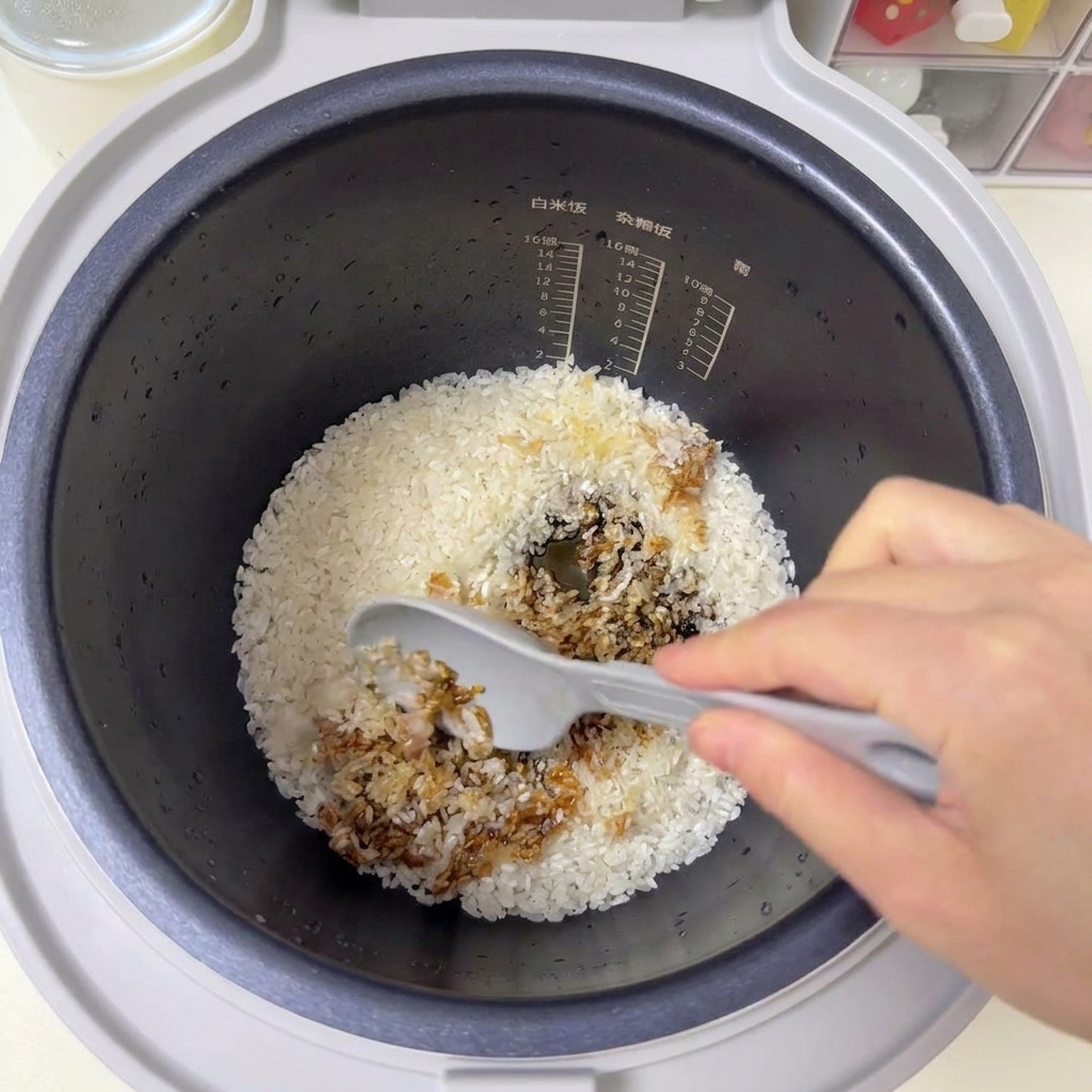 Una espátula gris mezclando arroz blanco y salsas oscuras dentro de una olla arrocera.