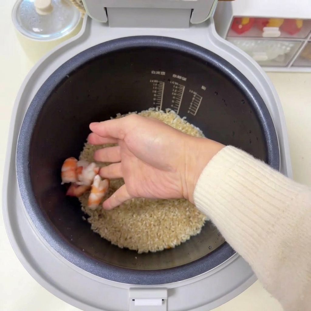Una mano colocando varios camarones rosados crudos sobre una cama de arroz sazonado en la arrocera.