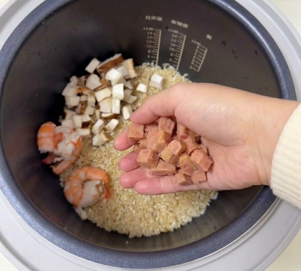 Una mano sosteniendo jamón picado en cubitos sobre la olla arrocera con arroz sazonado y camarones.
