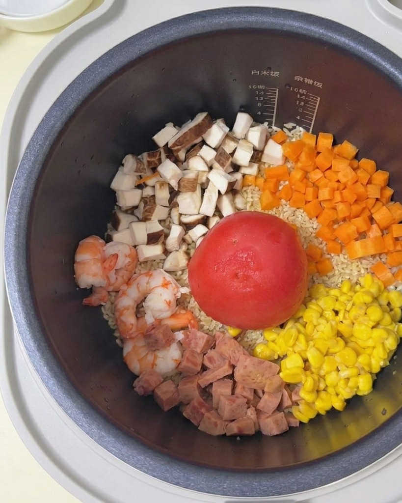 Agua vertiéndose desde un tazón blanco a la olla arrocera llena de arroz, tomate y diversos ingredientes.